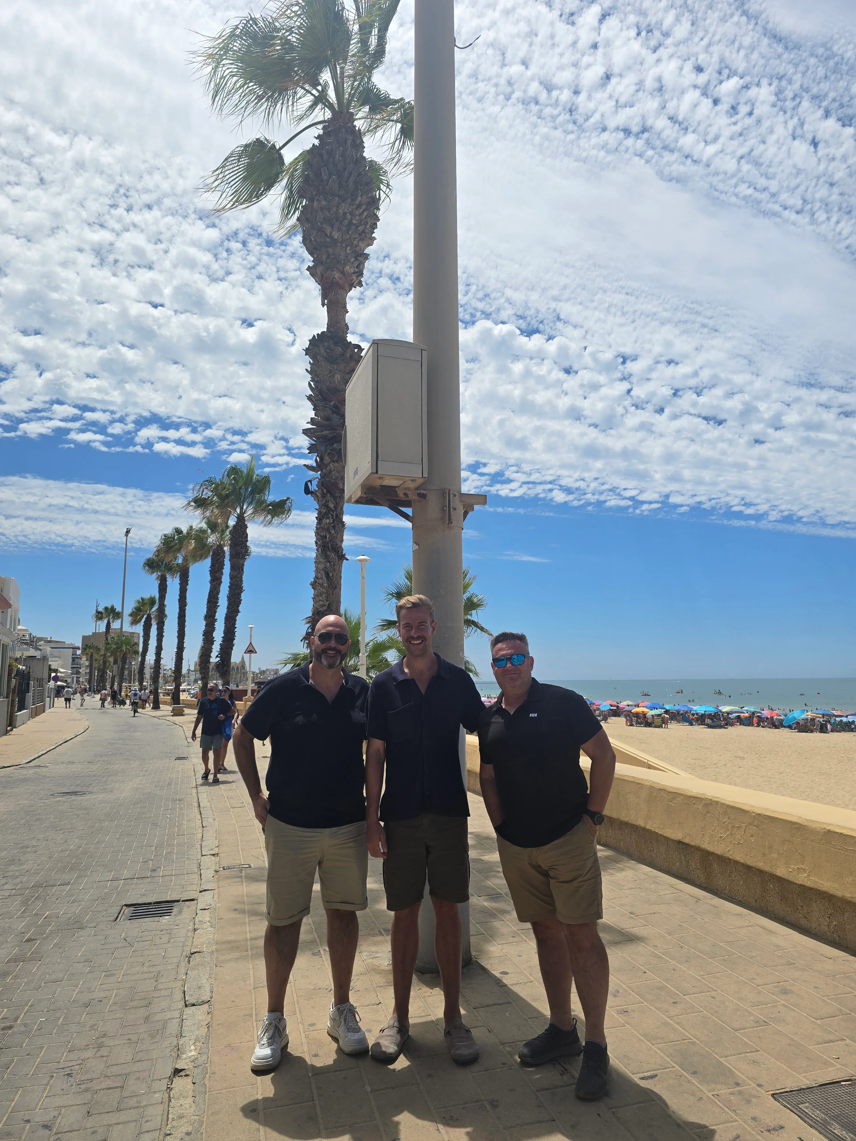 Alex Piatek, Pedro y Francisco Márquez (Paco) junto a la instalación de una cámara costera en el paseo de Chipiona, con la playa al fondo.